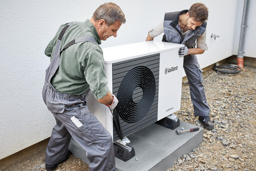 Zwei Monteure heben im Außenbereich eines Neubaus eine Wärmepumpe von Vaillant auf den Montage-Sockel. 