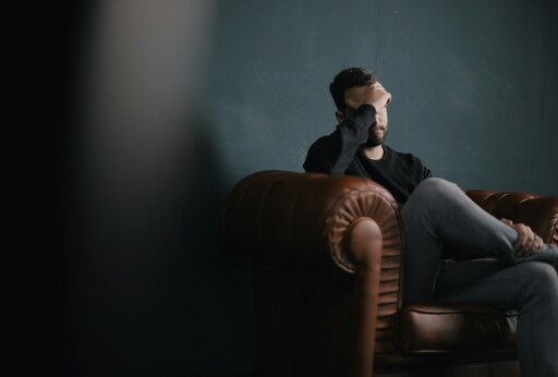 Ein trauriger Mann sitzt auf dem Sofa: Was passiert, wenn ein ein Partner plötzlich arbeitslos wird? Dann wird es auch mit der Kredittilgung eng.