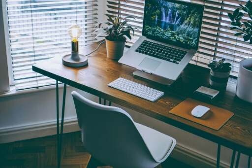 Ein Laptop auf Laptop-Ständer, davor eine Tastatur auf einem aufgeräumten Schreibtisch. Homeoffice vor heruntergelassenen Jalousien. 