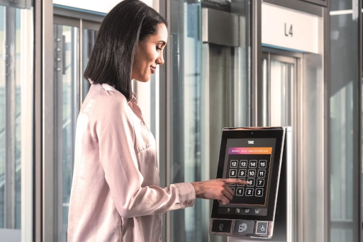 Eine Frau steht seitlich zum Aufzug  im 4. Stockwerk und drückt auf einem Touchpad die Taste 6. 