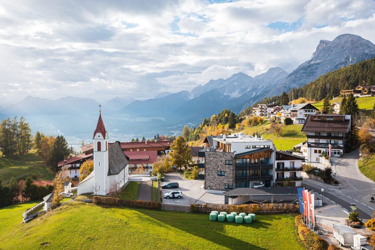 Eine ideale Umgebung für alle, die eine Ferienimmobilie suchen: Dieses Bild zeigt ein Dorf in Tirol. Im Vordergrund das von Sonnenstrahlen beleuchtete Neubauprojekt, im Hintergrund die Berge. 