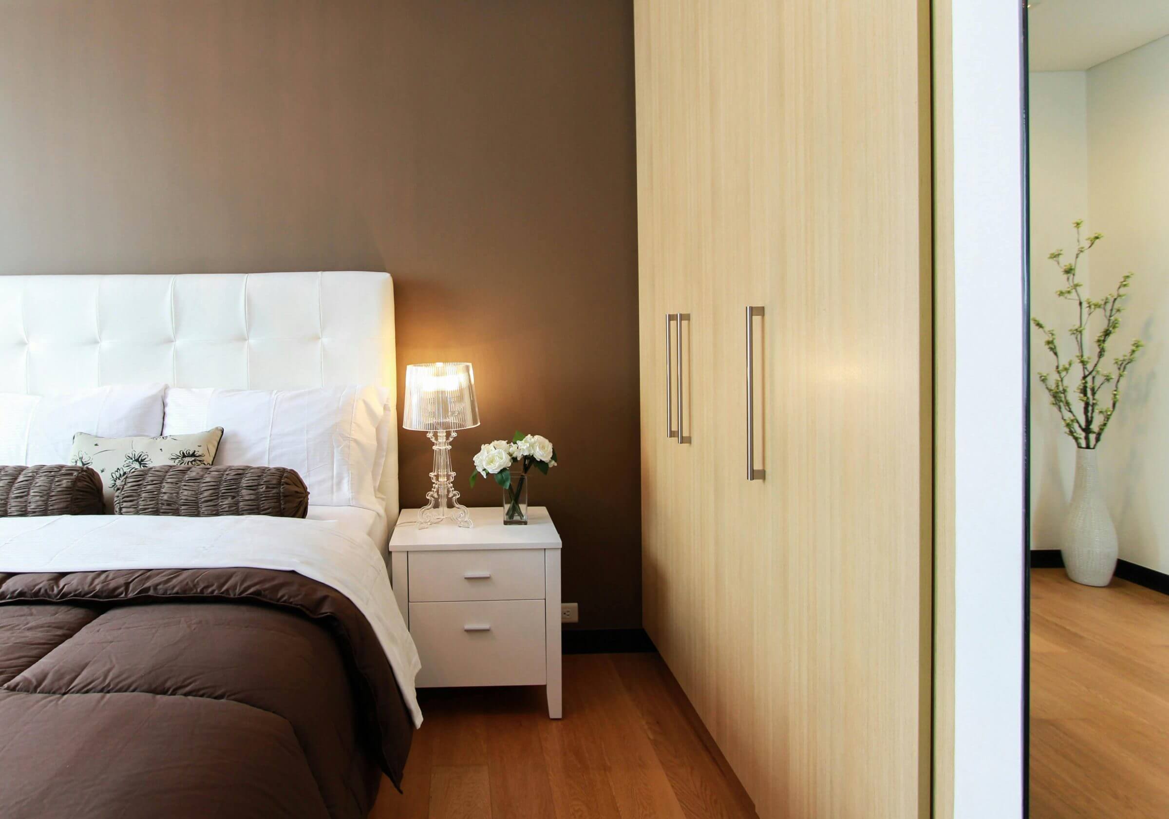 Gemütliches Schlafzimmer mit brauner Akzentwand, einem weißen gepolsterten Bettkopfteil, brauner Bettwäsche, einem weißen Nachttisch mit Lampe und Blumen sowie einem modernen Holz-Kleiderschrank.