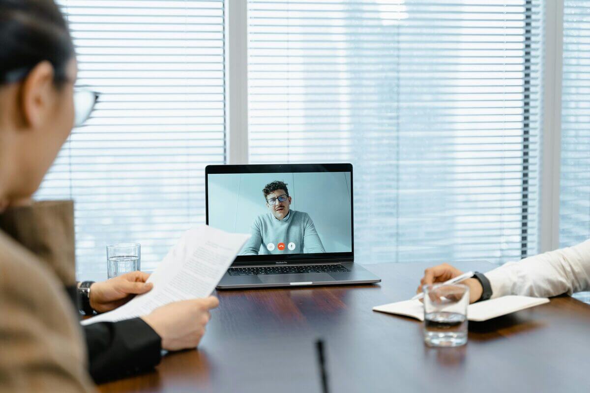 Videokonferenz: Mehrere Personen sitzen an einem Tisch vor einem Laptop in einem hellen Büroraum.
