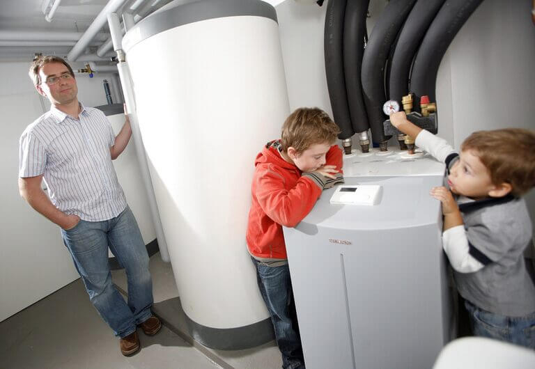 Ein Mann steht mit zwei Kindern um eine Wärmepumpe im Keller.